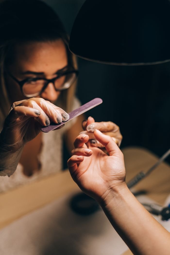 gallery-1 A detailed view of a manicurist filing a womans nails under soft lighting.